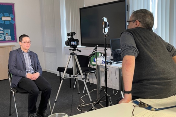 Young person being interviewed. Sitting on a chair in front of the camera, hands in lap.