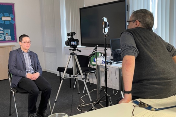 Young person being interviewed. Sitting on a chair in front of the camera, hands in lap. 