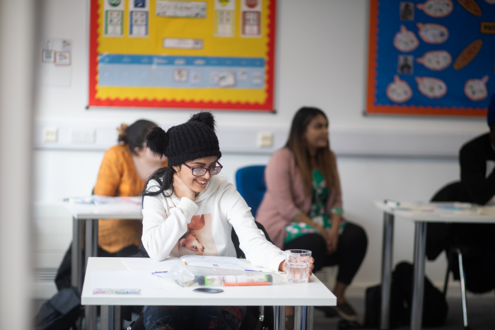 Young person sitting at desk in classroom, looking down at their work, smiling.