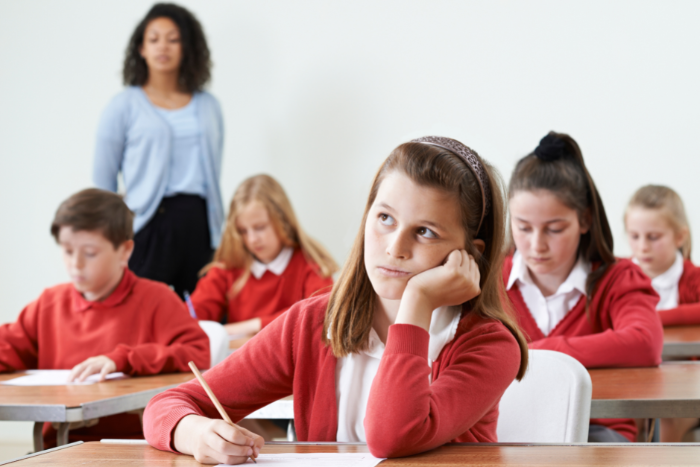 Pupil in school uniform, sitting at desk with exam paper in front of them. Teacher standing in background