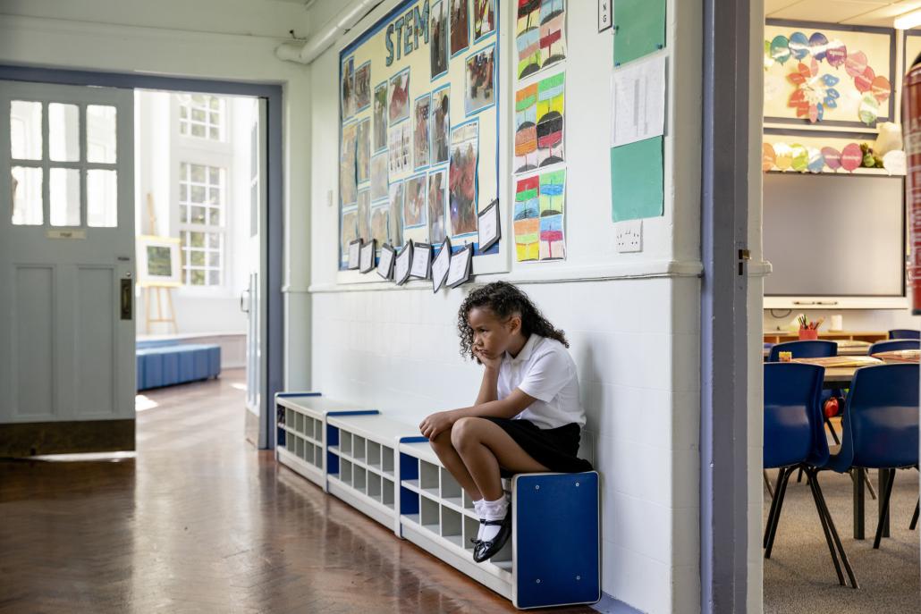 School pupil sitting on a bench outside a school classroom, their head in hand and looking at the floor. 