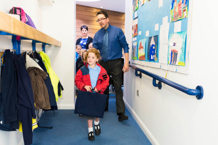 A primary aged pupil walks into the school corridor with their book bag and coat. A teacher is walking closely behind them.